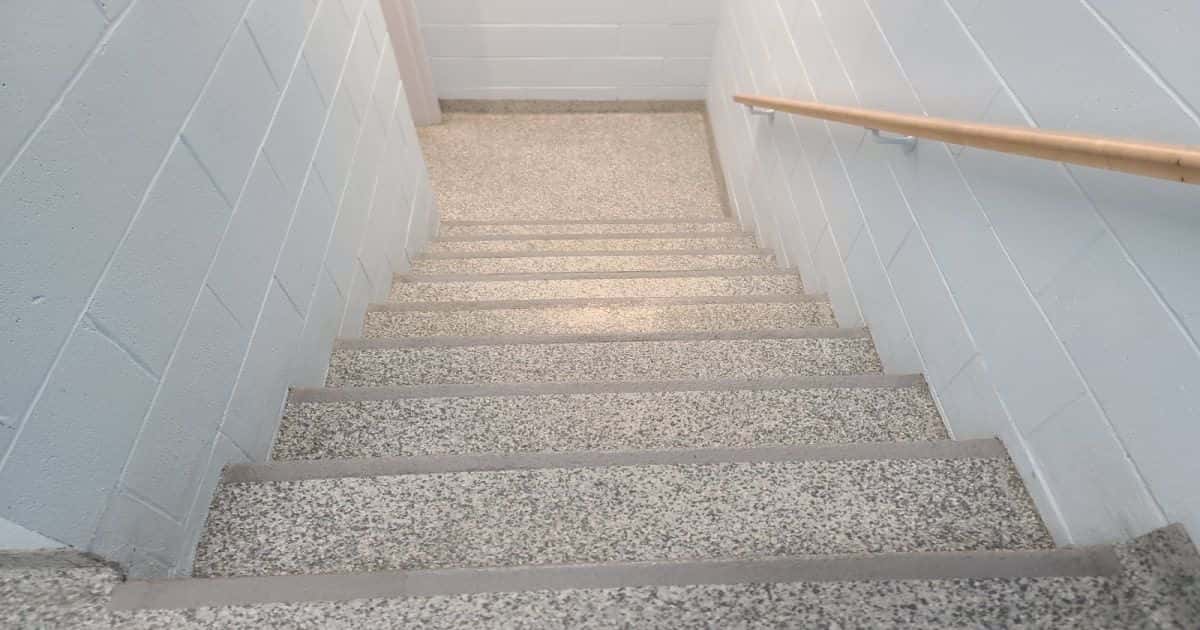 Een trap bekleed met siergrind en antislip per trede. Plus, een houten trapleuning die bevestigd is aan een witte muur.