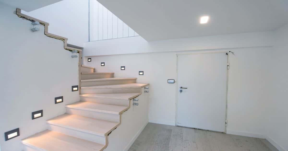Een trap met een combinatie van beige treden, witte muren en een grote trap gat. Er is verlichting in de muur langs elke trede. Er staat ook een grote witte deur op de foto.