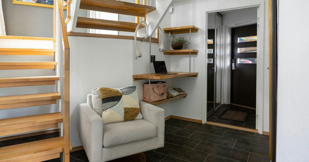 Gezellige zithoek onder een open, houten bordestrap op een zwarte tegelvloer die uitgeeft op de inkomhal. Aan de wand onder de trap hangen verschillende planken die als opbergruimte dienen. 