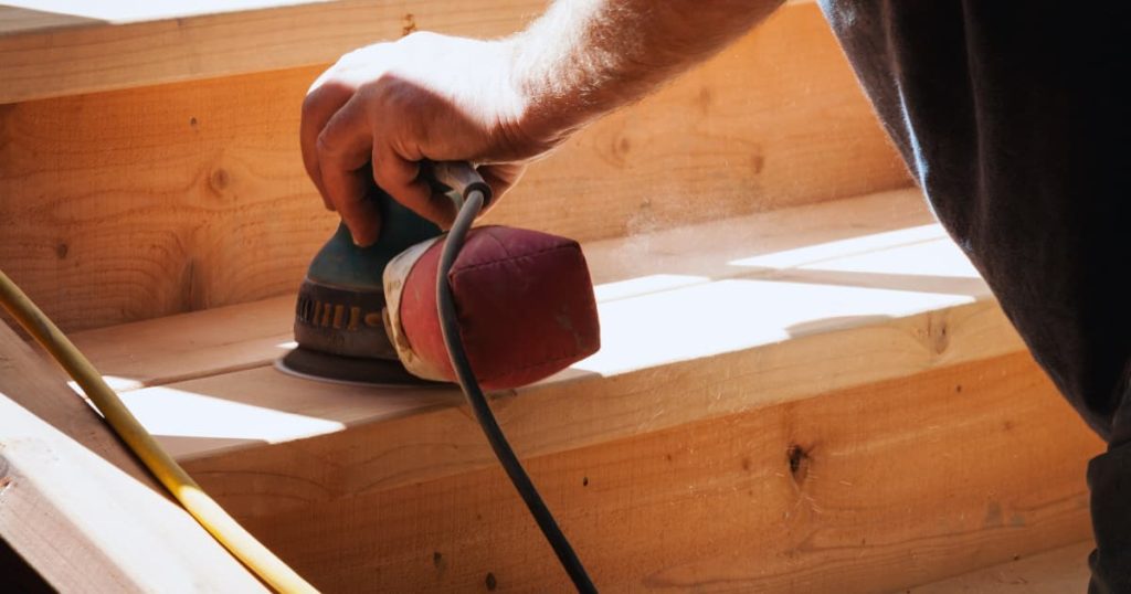 Dit is een mannenhand die met een schuurmachine de trap schuurt.