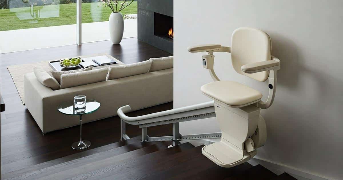 Een korte, donkere houten trap met witte stoellift. De trap geeft uit op een benedenverdieping met donkere houten vloer, grote glazen wanden, en een modern interieur met witte zetel, minimalistische openhaard in zwarte wand, en grote vaas.