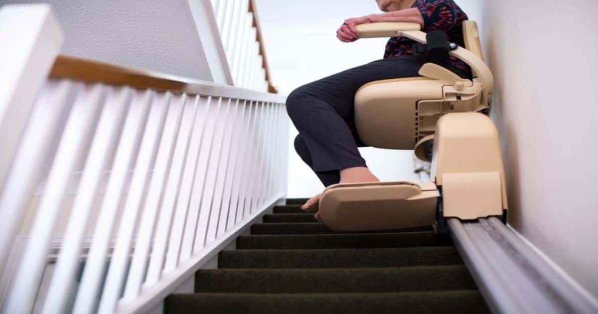 Een traplift die een steile trap opgaat, een oude vrouw zit op de traplift.