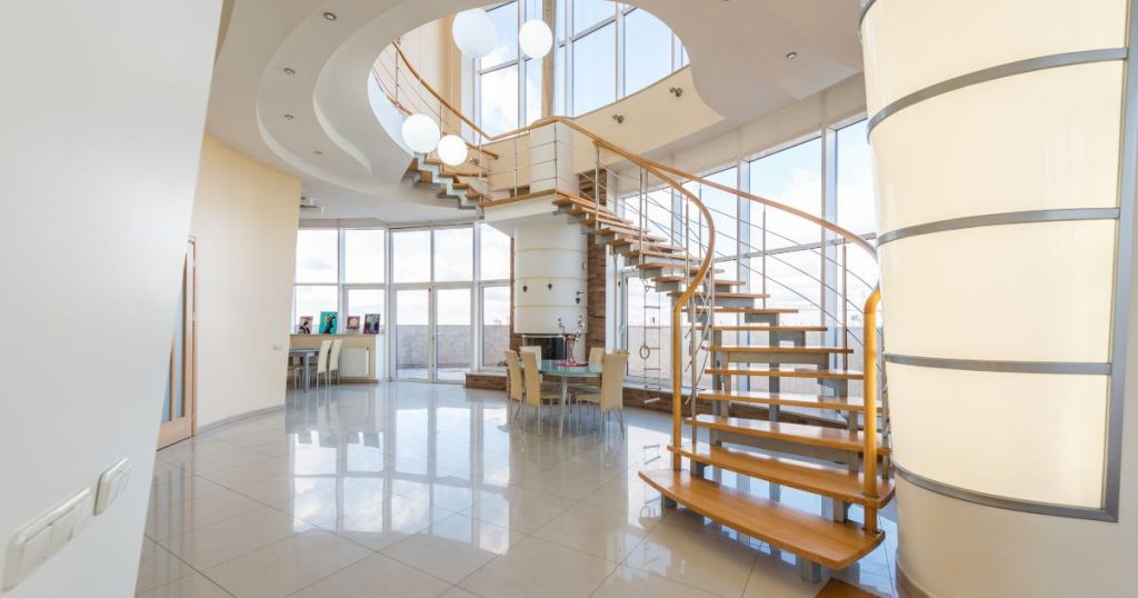 Een houten open wenteltrap in een moderne woning. De trap begint op een glanzende tegelvloer, loopt via een bordes omhoog en mondt uit in een ruim, modern rond trapgat. 