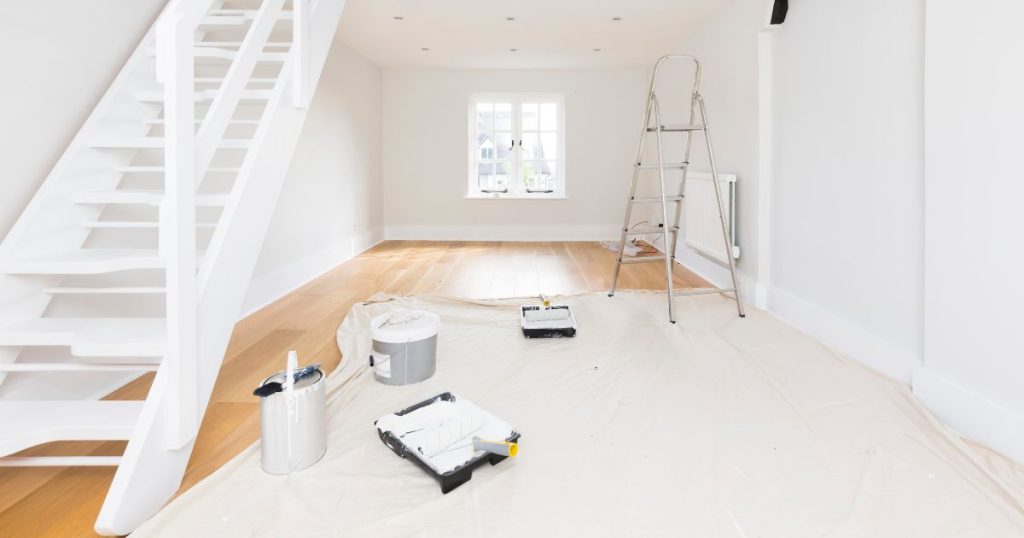 Een kamer met houten vloer, witte muren en een witte molenaarstrap of open houten steektrap. In de ruimte ligt een zeil om de vloer te beschermen tijdens het schilderen. Op het zeil staat een trapladder, twee verfbakjes met verfrollers, twee emmers verf en een verfkwast.