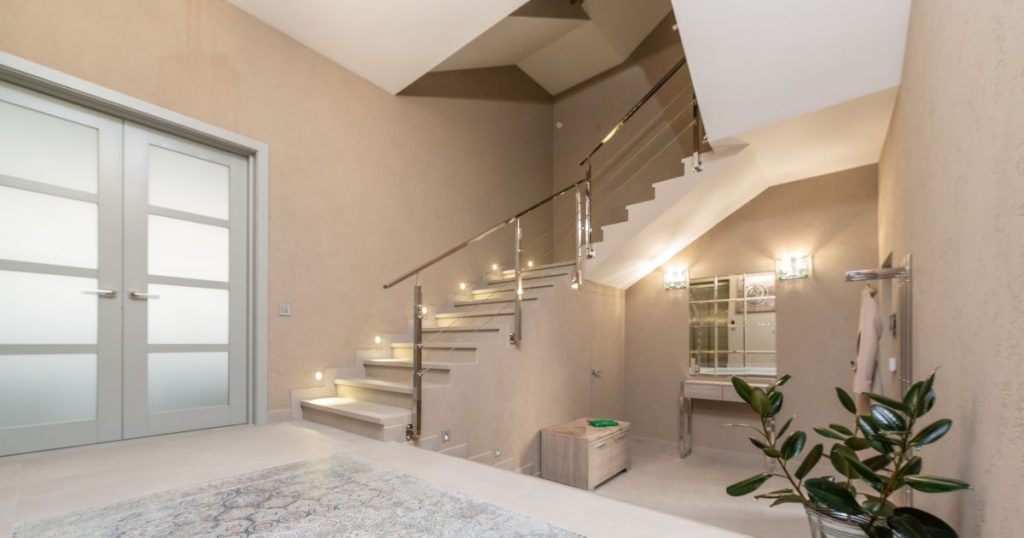 Een statige trap in een lichtroze tint met trapverlichting, ijzeren trapleuning en antislip strips in een bruinroze trappenhal met spiegel, plant, tapijt en dubbele deur met verticale, opake ramen.