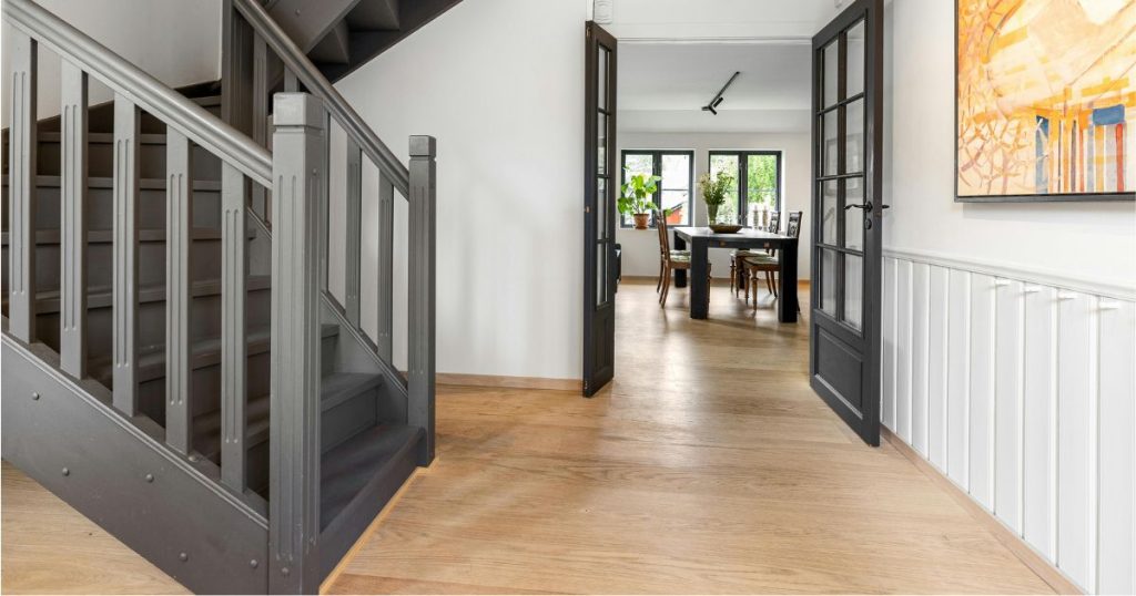 Een trappenhal in een woning met een rustig interieur en houten vloeren. In de hal staat een zwarte kwartslagtrap, en een geopende zwarte houten deur met ramen geeft uit op een harmonieuze eetkamer met grote eettafel met vier stoelen.