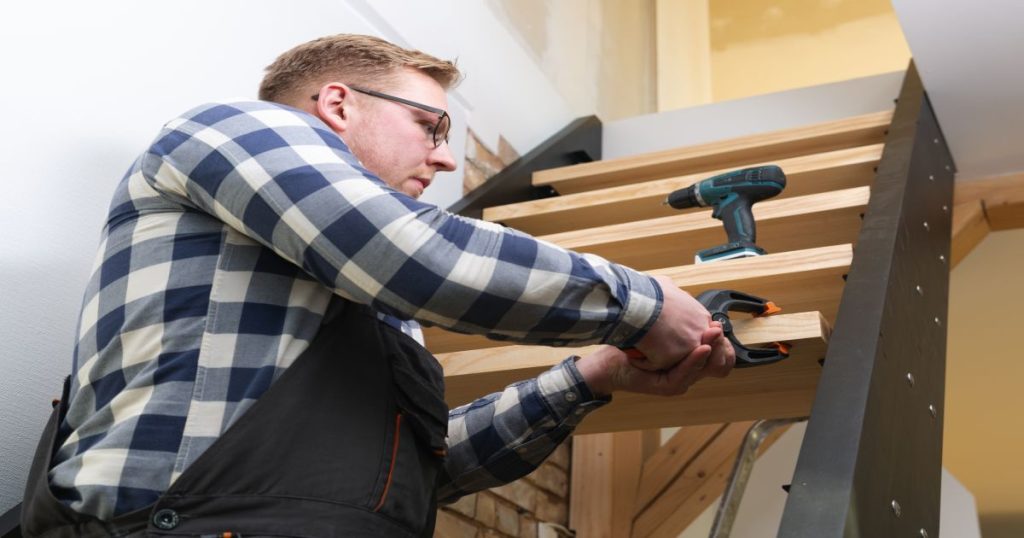 Een man die zijn eigen trap renoveert. Hij houdt met een klem een traptrede vast die hij voorzichtig probeert te plaatsen.