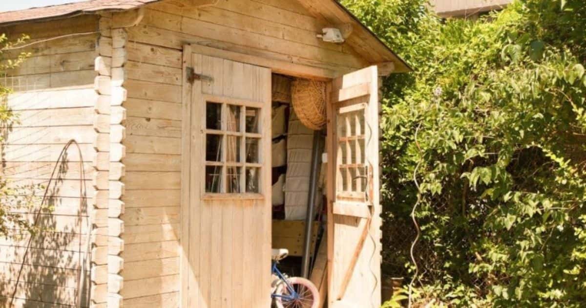 Tuinhuis in licht hout: de deur staat open. Je kan een rieten mand zien, een het frame en achterwiel van een blauwe fiets, een grijze paal of stok, en beige zakken die ophangen. Rond het tuinhuis staat veel dichte begroeiing.