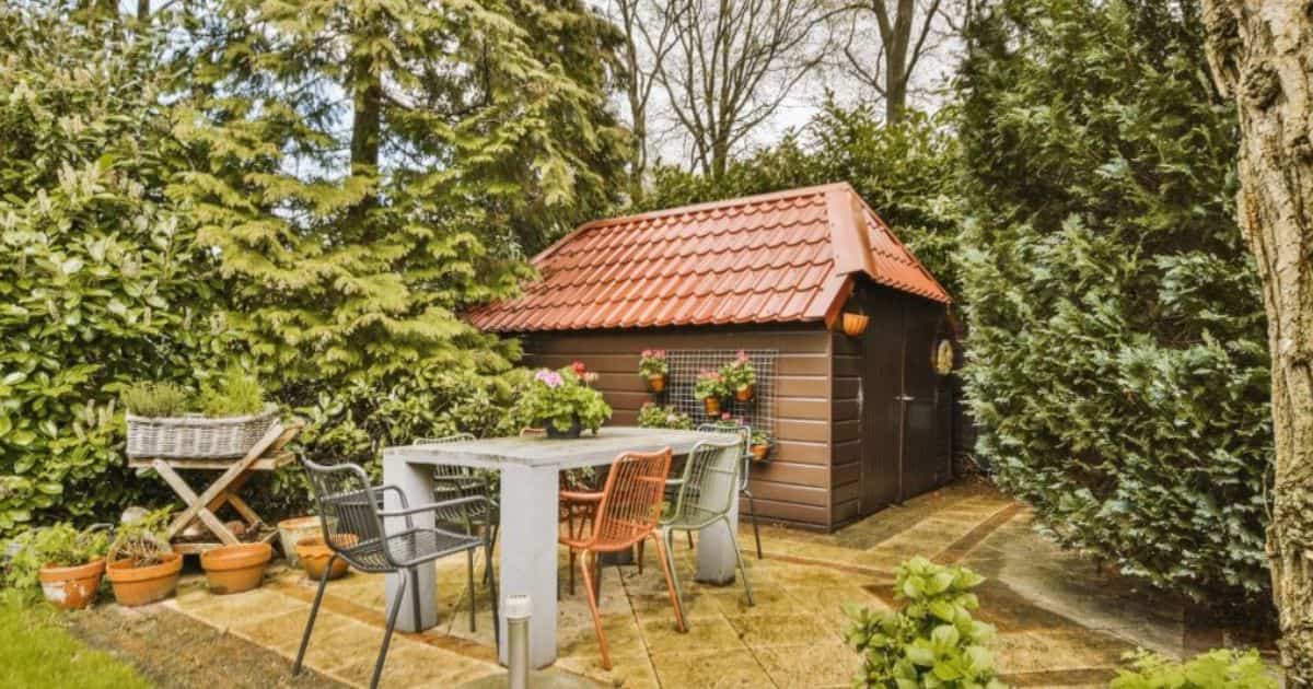 Tuinhuis met wolfsdak met oranje dakpannen. Tegen de gevel hangen bloempotten met rode bloemen.