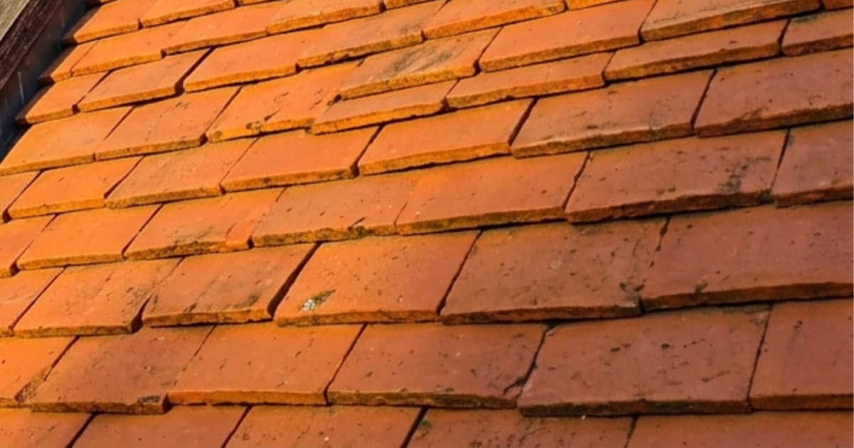 Twee snijdende hellende daksecties met platte tegelpannen in roodbruine en oranjebruine tinten gescheiden door een diagonale zinken kilgoot tegen een heldere lucht.