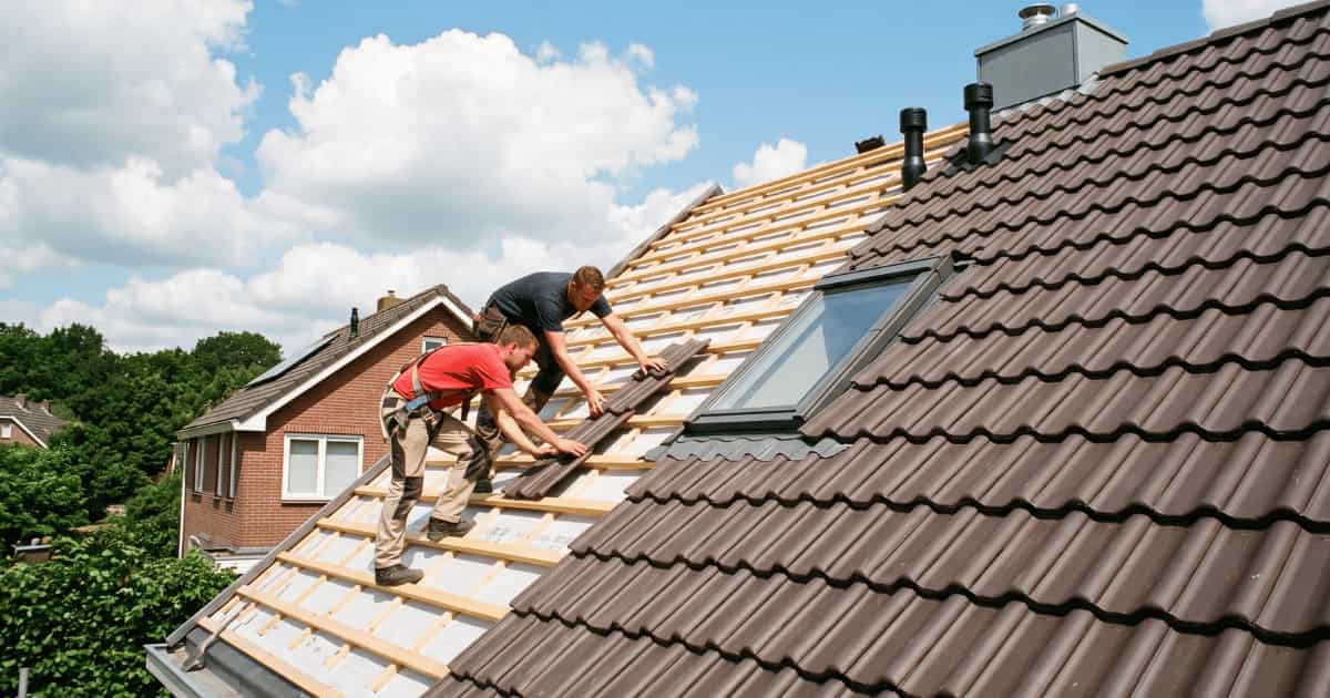Twee dakwerkers plaatsen bruine betonnen sneldekpannen op houten panlatten aan de linkerzijde van een hellend dak terwijl de rechterzijde al volledig betegeld is met een dakraam ertussen en zwarte ventilatiepijpen bij de nok tegen een blauwe lucht met witte wolken.