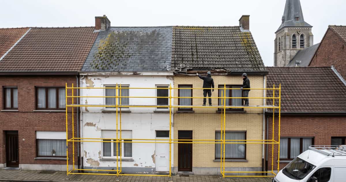 Twee dakwerkers in donkere werkkleding en helmen staan op een gele stelling ter hoogte van de dakrand van twee aanpalende Belgische rijwoningen met een verweerd leistenen dak vol mos, een afbladderende witte gevel links en een lichtgele bakstenen gevel rechts, met aangrenzende rode bakstenen rijwoningen en een kerktoren op de achtergrond onder een bewolkte hemel.