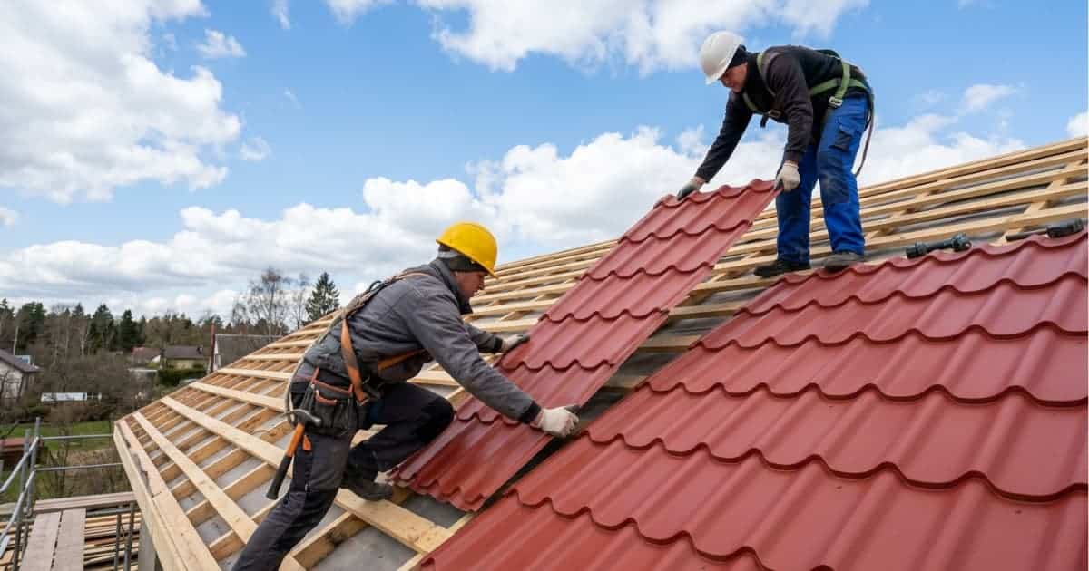 Twee dakwerkers met veiligheidshelmen op een hellend dak in aanbouw grijpen samen een grote rode metalen dakpanplaat met gestanst golfpatroon en schuiven deze in positie naast reeds geplaatste platen, met onbedekte houten panlatten zichtbaar en een blauwe lucht met witte wolken op de achtergrond.