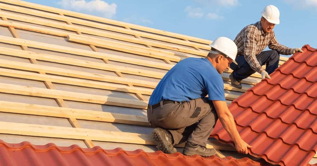 Twee dakwerkers met witte veiligheidshelmen hurken op een hellend dak en positioneren samen een grote rode metalen dakpanplaat op verse houten daklatten terwijl het lagere gedeelte al bedekt is met identieke rode panplaten onder een blauwe lucht.