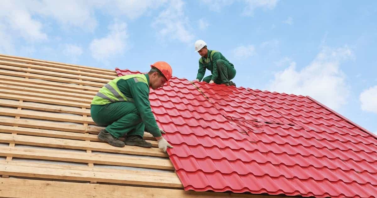 Twee dakwerkers in groene werkkleding plaatsen rode geprofileerde metalen dakplaten op een hellend dak, de onderste werker met oranje helm en fluogeel hesje hurkt op de blote houten panlatten en schuift een plaat op zijn plaats terwijl de bovenste werker met witte helm bij de nok op de reeds gelegde platen zit, tegen een blauwe lucht met witte wolken.