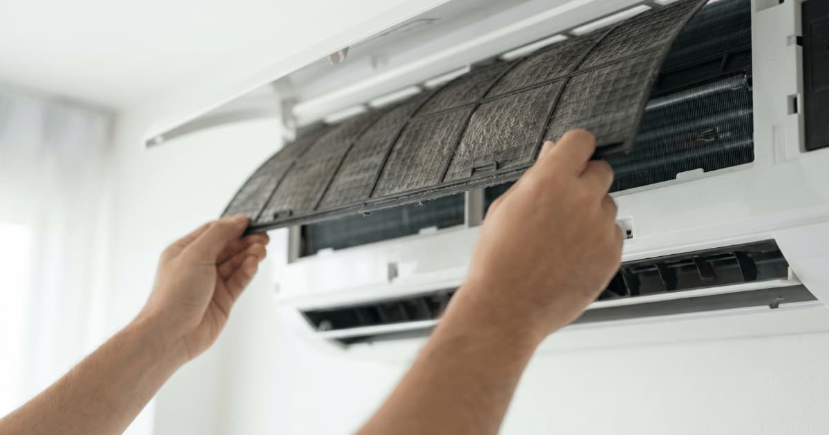 Twee handen grijpen de randen van een zichtbaar vervuild donker filter en schuiven het uit de geopende witte airco-binnenunit terwijl de zwarte verdamperlamellen erachter zichtbaar worden.