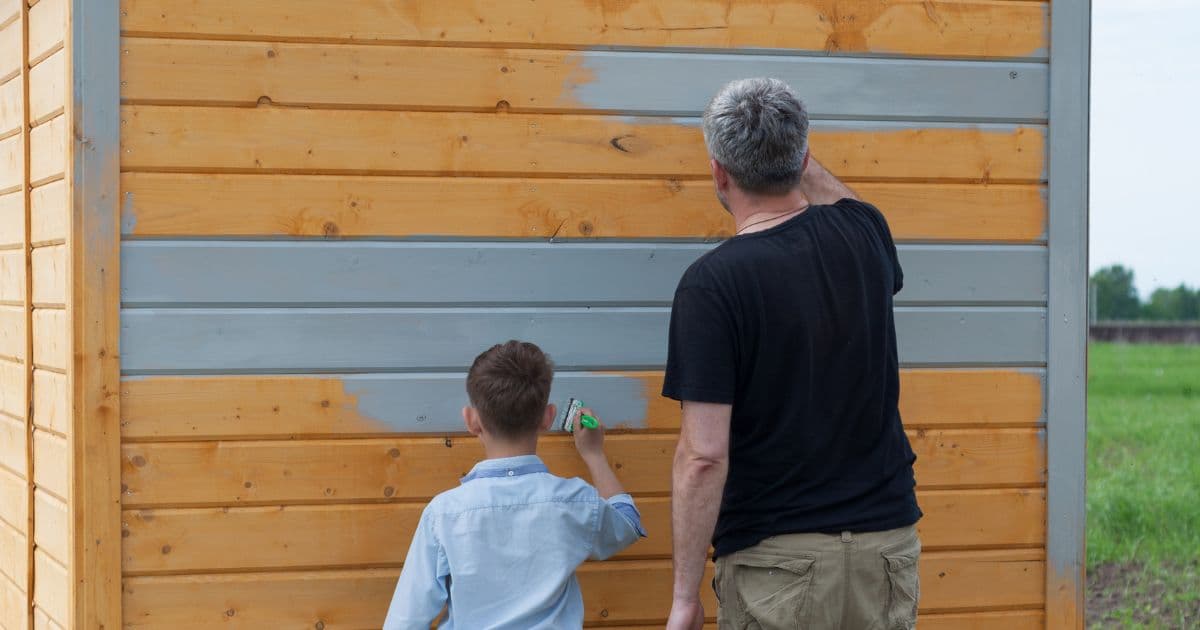 Een vader en zoon verven samen een houten tuinhuis grijs. De vader schildert de bovenste planken, terwijl de zoon de onderste voor zijn rekening neemt.