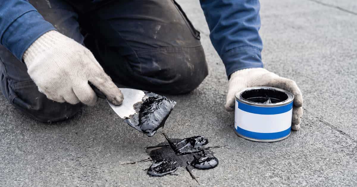 Een vakman met witte werkhandschoenen knielt op een grijs plat dakoppervlak en brengt met een plamuurmes zwarte bitumen herstellingspasta aan over een scheur in de dakbedekking terwijl zijn andere hand een geopend blik met dezelfde pasta vasthoudt.