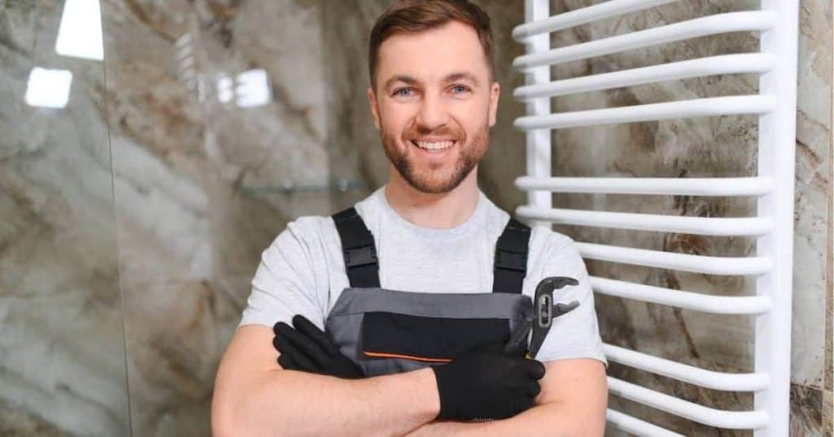 Lachende vakman met werkhandschoenen en overall, met de armen gekruist, in een badkamer met marmerlook-tegels en een witte handdoekradiator.