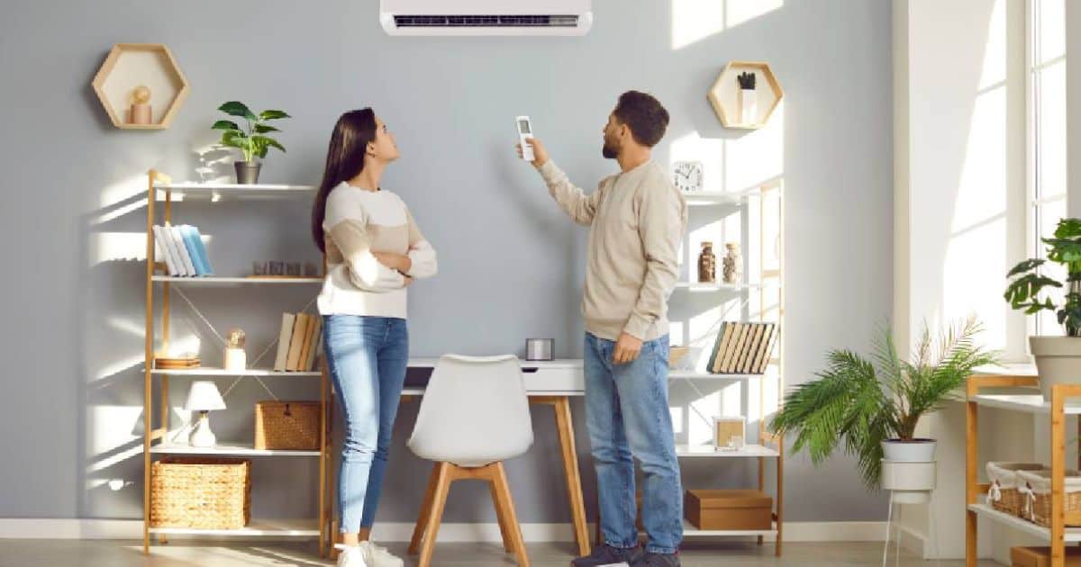 Een koppel staat in een lichte living. De man gebruikt een afstandsbediening om de temperatuur te regelen met de binnenunit van een split airco systeem dat zowel kan koelen als verwarmen.