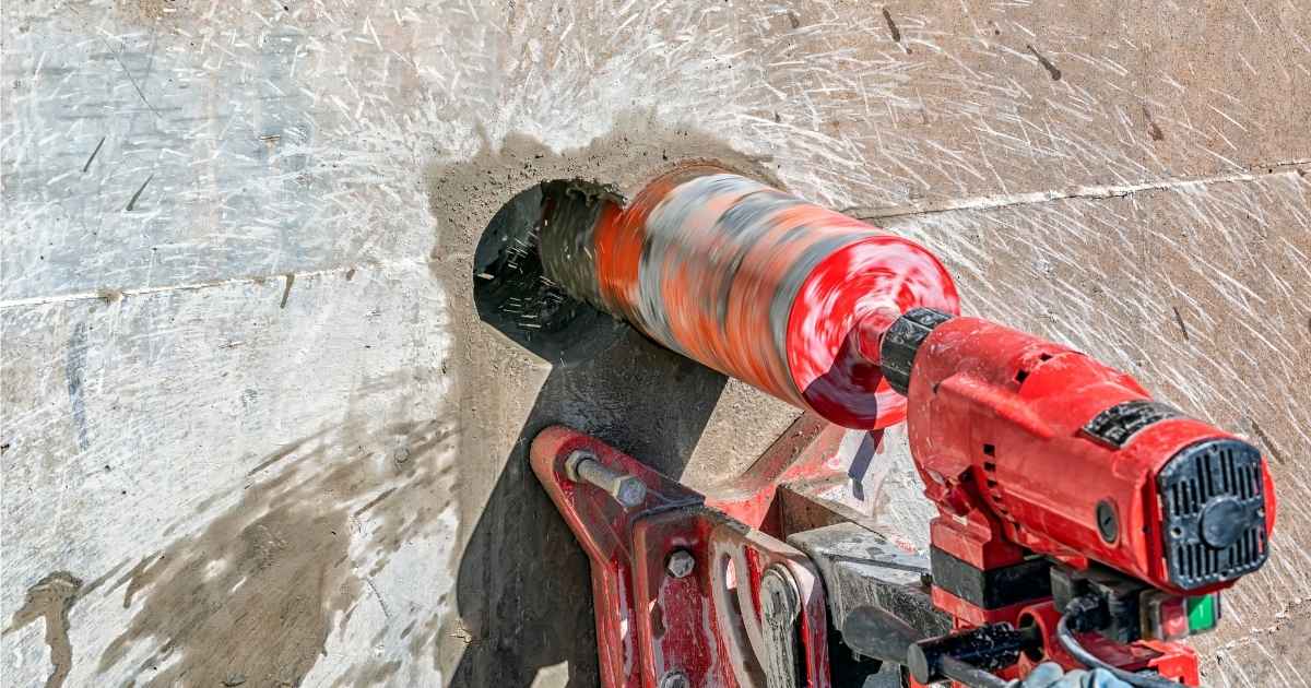 Een rode betonboor tegen een betonnen muur. In de wand zit een relatief oppervlakkig gat met een diameter van enkele centimeters.
