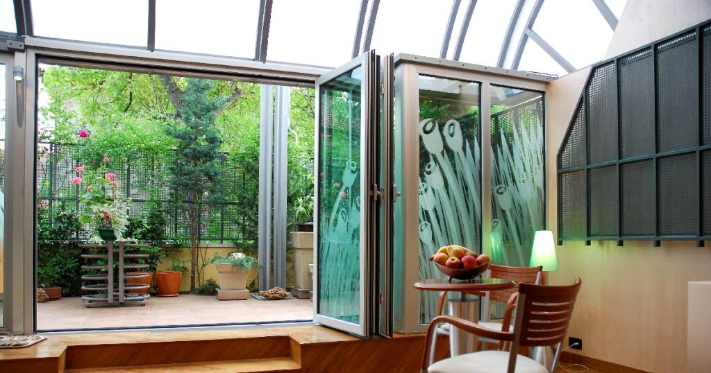 Een kleine veranda met transparant dak uit polycarbonaat dat moeilijk te isoleren valt. De veranda geeft uit op een kleine tuin via een grote dubbele zwaaideur.