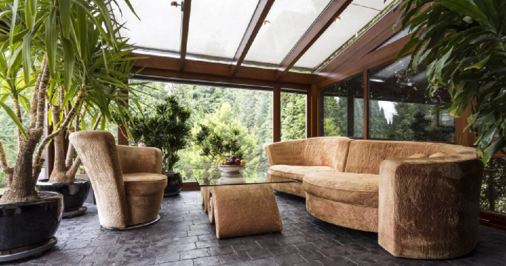 Serre met veel natuurlijk licht. De serre is van glas en hout gemaakt. In de serre staat een bank met een stoel, bijzettafel en een aantal planten.