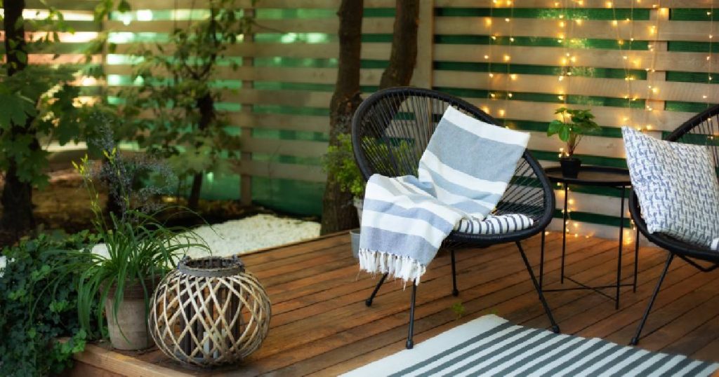 Een houten veranda, dichtgemaakt met houten sidings en groene doeken. In de veranda staan zwarte kuipstoelen, een bijzettafeltje, een blauw met wit gestreept matje, enkele kamerplanten en een decoratieve bol.