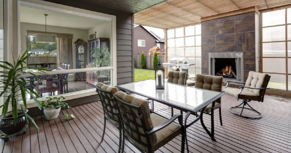 Een half open veranda tegen een woning. Men heeft de veranda deels laten dichtmaken met mat glas in een houten frame. De veranda heeft een donker houten vloer. Tussen twee glazen wanden (vaste kozijnen) is een openhaard geplaatst. In de veranda staan een glazen tafel, 5 stoelen en verschillende kamerplanten. Links de living van de woning, zichtbaar door een groot raam met wit frame in een gevel van donker hout.