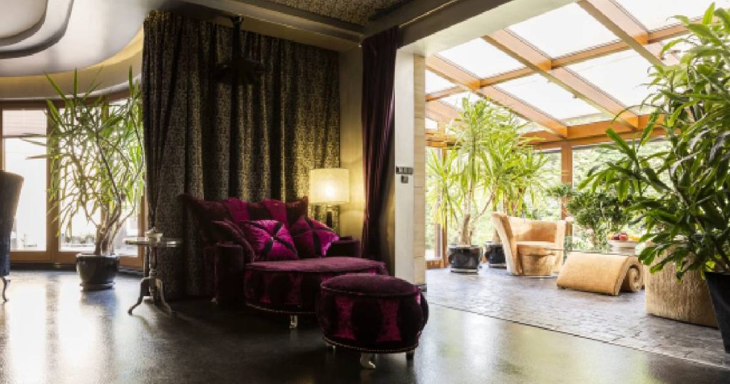 Een luxe woning met een volledig glazen veranda. De woning heeft een donkere vloer die uitloopt op de lichte tegelvloer van de veranda. In de woning staat een paarse zetel met poef, in de veranda lichtgekleurde zetels, grote kamerplanten en een glazen tafel met stoffen steun.
