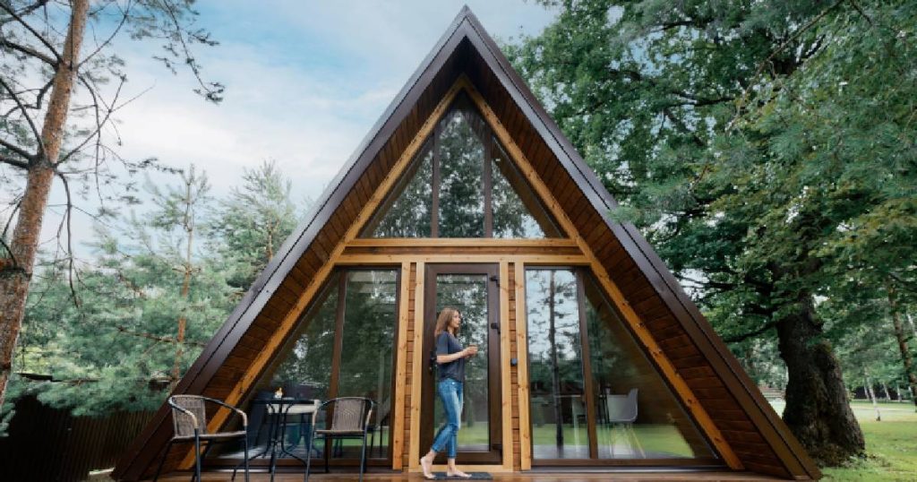 Een vierkante, losstaande veranda uit hout en glas. Voor de veranda loopt een dame in blauwe jeansbroek en zwart t-shirt. Voor de veranda staat een tafeltje met twee tuinstoelen. Achter de veranda bomen en gras.