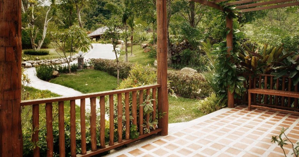 Een veranda die men deels heeft laten dichtmaken met een lage, open schutting uit hout. De veranda geeft uit op een weelderige tuin waar een wit pad door kronkelt. De veranda staat op een vetegelde vloer.