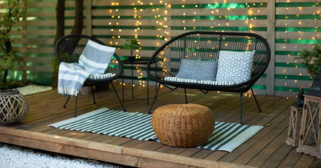 Een volledig houten veranda die men heeft laten dichtmaken met planken. Op de houten verandavloer staan een grote en kleine zetel met dekens en kussentjes. Er ligt ook een gestreept matje op de vloer.