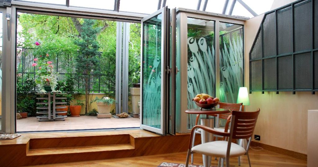 Een gezellige veranda die men deels heeft laten dichtmaken met plexiglas. De houten vloer loopt over in een betegeld terras. In de veranda staat een tafeltje met twee stoelen en een fruitmand.