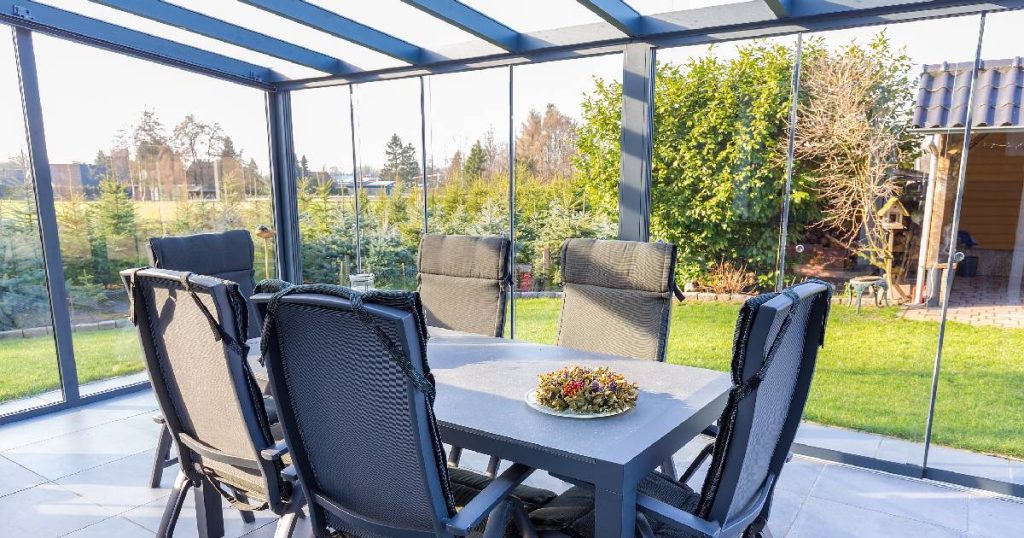Een mogelijke uitvoering van je veranda dichtmaken met plexiglas. In de veranda of serre staat een buitentafel met stoelen. De veranda geeft uit op een grasveld omringd met struikgewas. Rechts in de hoek is een stuk van een half open tuinhuisje zichtbaar.
