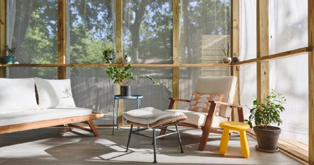 Een veranda die de eigenaars hebben laten dichtmaken met screens en houten latjes. In de veranda staan houten zetels met witte kussens, een bijzettafeltje, een voetenbankje, een geel plastic krukje en twee kamerplanten.