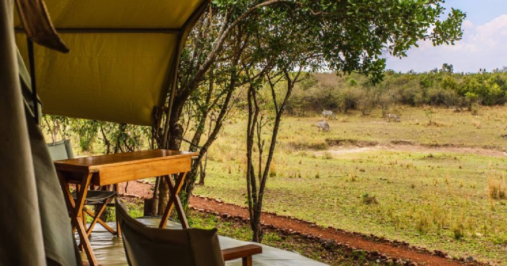 Zijaanzicht van een veranda die de eigenaars hebben laten dichtmaken met hout en een canvas zeil. De veranda geeft uit op een open vlakte. Op de houten verandavloer staan houten klapstoeltjes met groene canvas zittingen en een houten tafel.