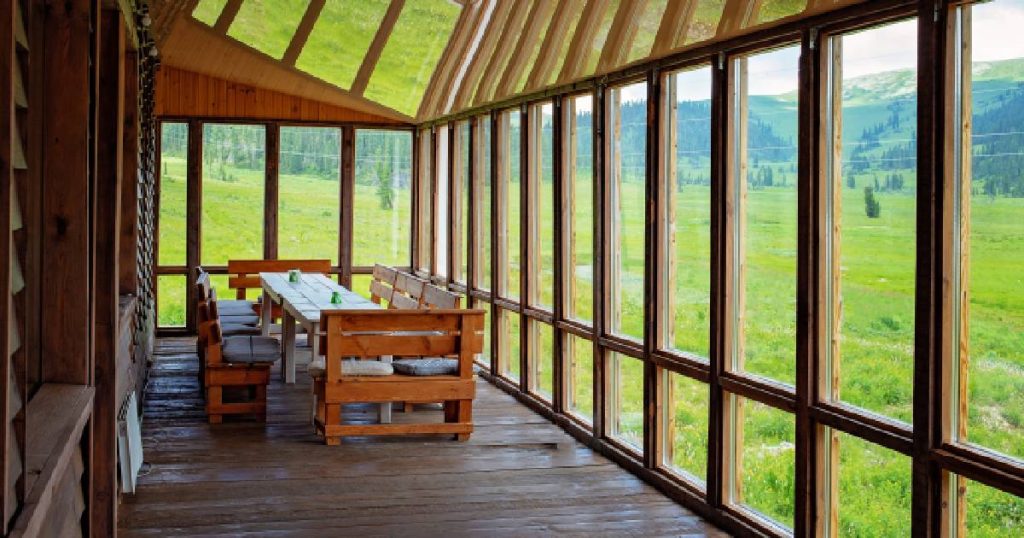 Een gezellige veranda in verschillende materialen, waaronder hout en glas. In de veranda ligt een houten vloer en staat een lange houten tafel met houten zitbankjes. De veranda geeft uit op een groene weide.