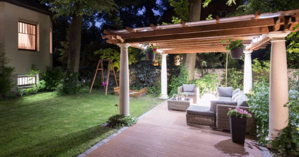 Een pergola gebouwd door een professionele verandabouwer. Onder deze open veranda met houten dak, houten vloer en steunpilaren staat een loungeset en brandt licht.