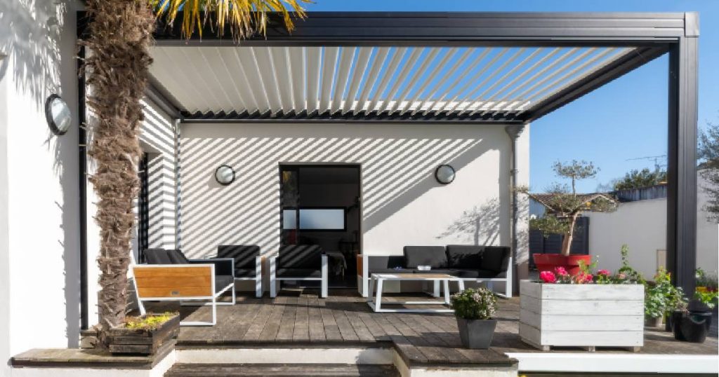 Een moderne, strakke pergola met lamellendak en aluminium draagconstructie, tegen een witte gevel gebouwd door een verandabouwer. De veranda heeft een houten vloer. In de veranda staan verschillende moderne tuinmeubels met zwarte kussens.