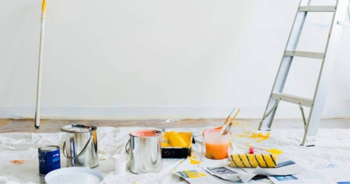 Een reeks verfpotten, een verfbakje met gele verf, borstels en een ladder op een wit doek op de grond. Alles staat klaar om de ruimte professioneel te schilderen.