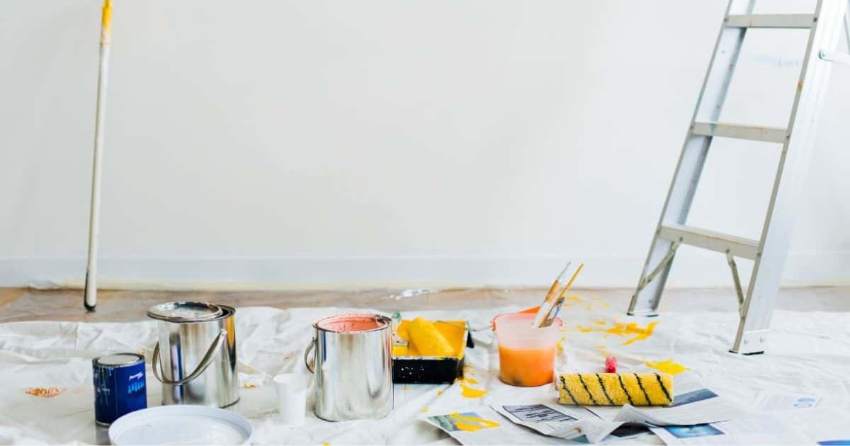 Een ruimte met witte muur, een ladder en een aantal verfpotten: één van de potten is geopend. Ze bevat een roze verfkleur. In het verfbakje zit een gele verfkleur. De borstels zitten in een pot met water dat de verfkleuren heeft overgenomen.