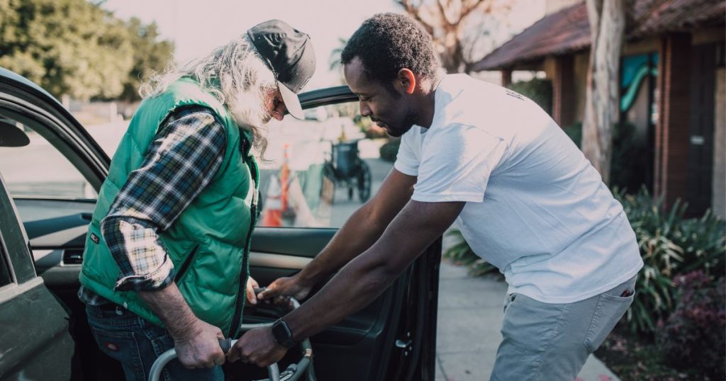 Een jonge man in wit t-shirt en jeansbroek helpt een ouder persoon met rollator, pet, groene bodywarmer en ruitjeshemd uitstappen uit een auto.