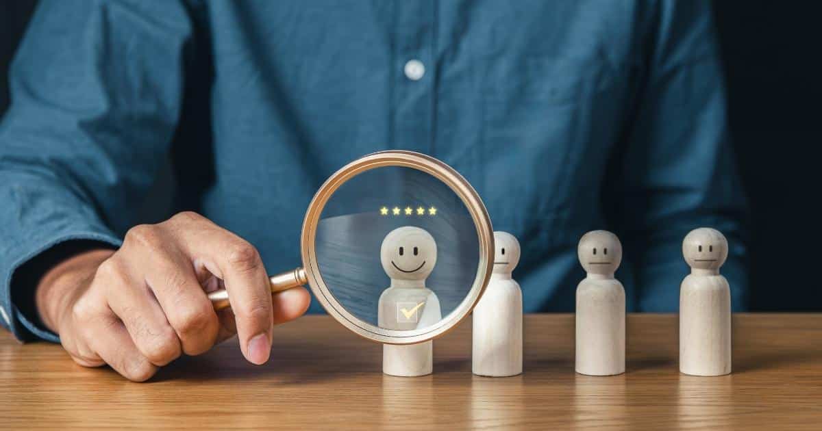Een man in blauw hemd houdt een vergrootglas voor een houten poppetje. Het poppetje heeft een glimlachend gezicht. In het vergrootglas zie je 5 sterren boven het hoofd van het poppetje, en een vinkje op de borst van het poppetje. Rechts van het poppetje met de glimlach staan nog 3 poppetjes met neutrale gezichtjes. Alle poppetjes staan op een houten tafel.