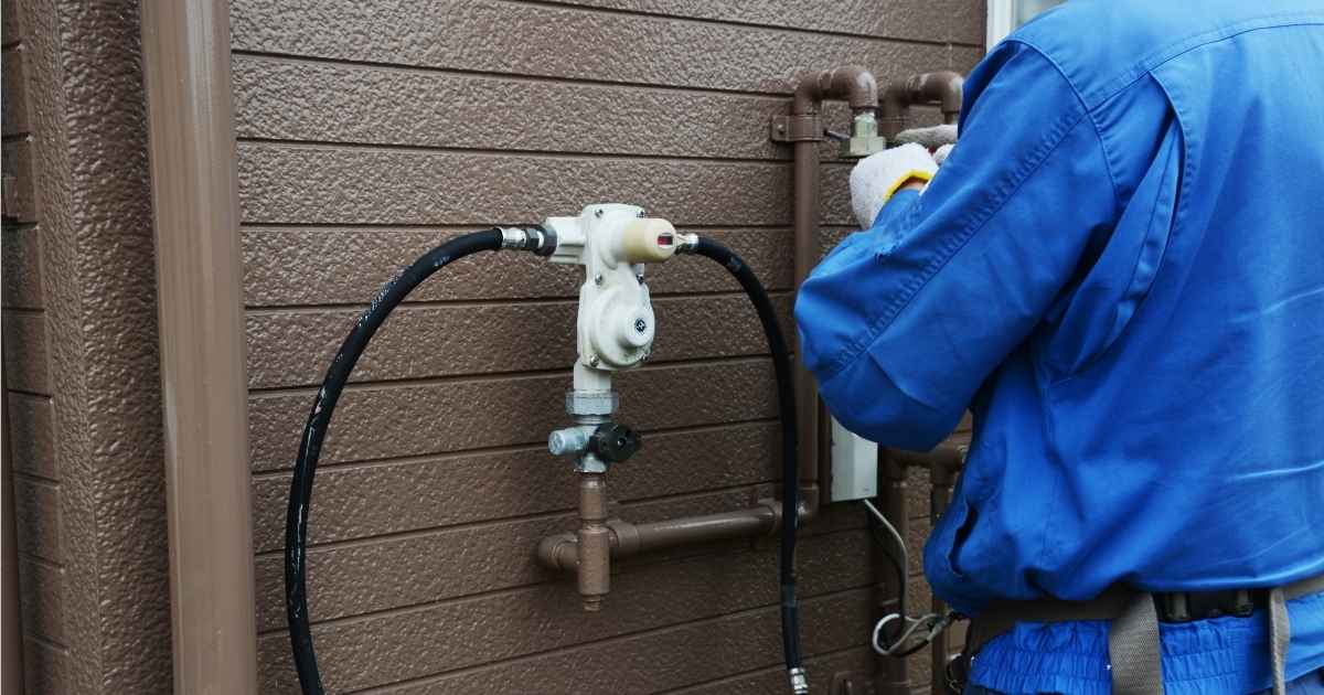 Een installateur in blauw uniform sluit een gastank aan op een woning met bruine kunststof gevel.