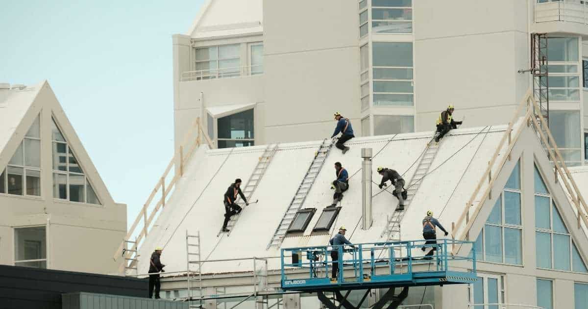 Vijf dakwerkers werken aan een nieuwe verdieping op een verhoogd hellend dak. De woning en de omliggende gebouwen zijn volledig wit.