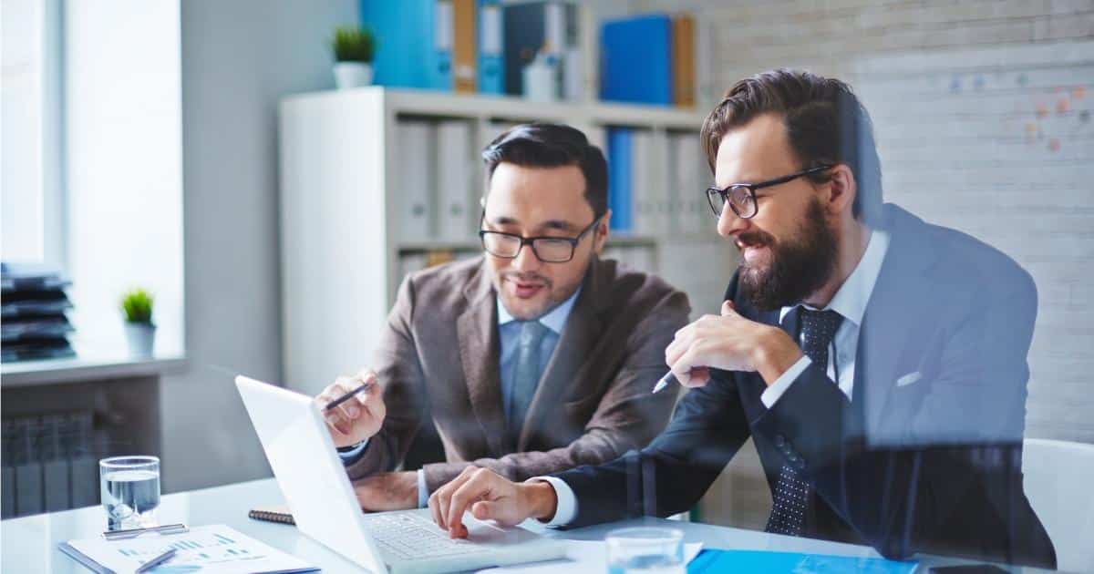 2 mannen in maatpak kijken samen naar een laptopscherm: de ene wijst met een balpen of potlood naar het scherm, terwijl de andere zijn hand op het toetsenbord richt. Op de tafel liggen ook documenten en staan 2 glazen water.