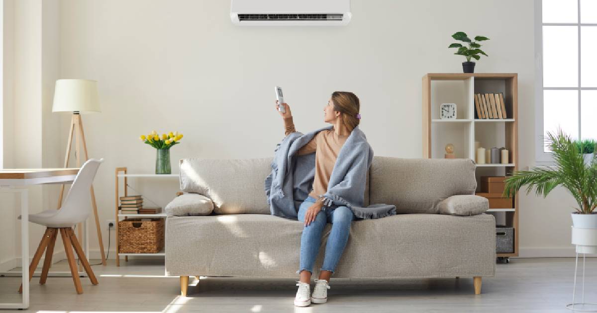 Een vrouw zit op een bank in een woonkamer onder een vaste airco die aan de muur hangt. Ze heeft een afstandsbediening vast om de airco mee te bedienen.