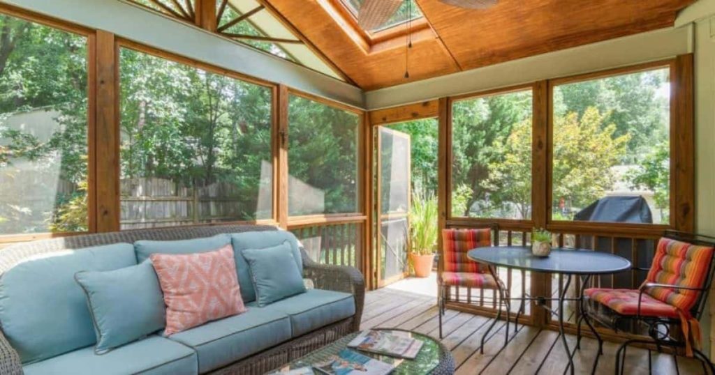 Gezellige houten veranda die als aanbouw tegen een woning werd geplaatst. Een houten vloer, grote raampartijen, gezellige zetel en tafel met twee stoeltjes met kussens maken de veranda een gezellige zitplek.