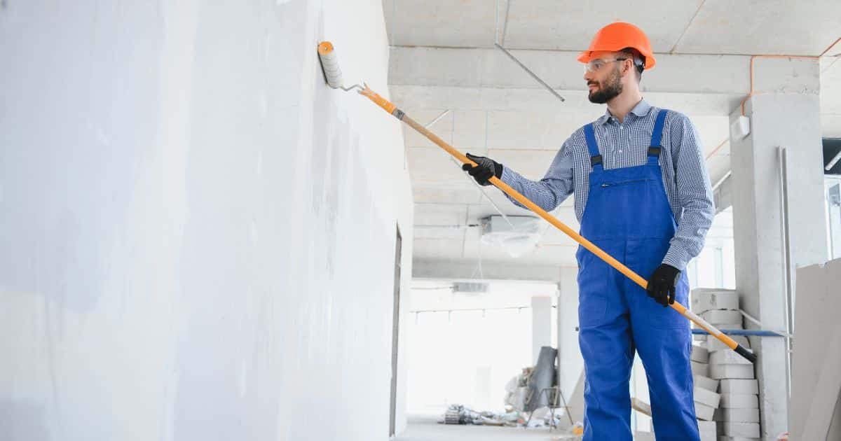 Een schilder in blauwe overall en met oranje helm schildert een muur in een woning.